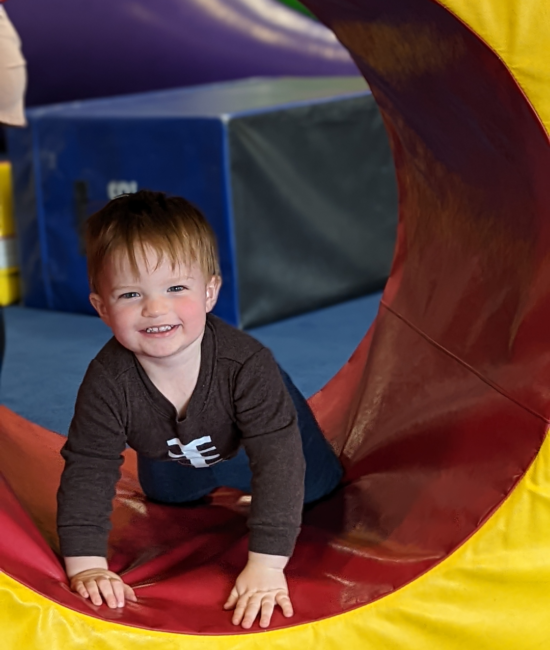 A smiling young child exploring a colorful play tunnel at Momentum